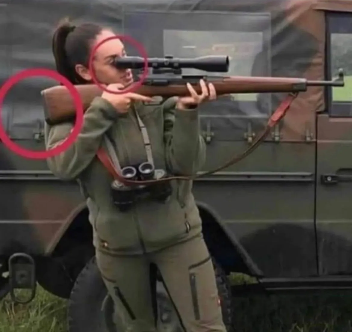 a woman in military-style green clothing holds a scoped rifle, aiming it with both hands. Her face is partially obscured by…