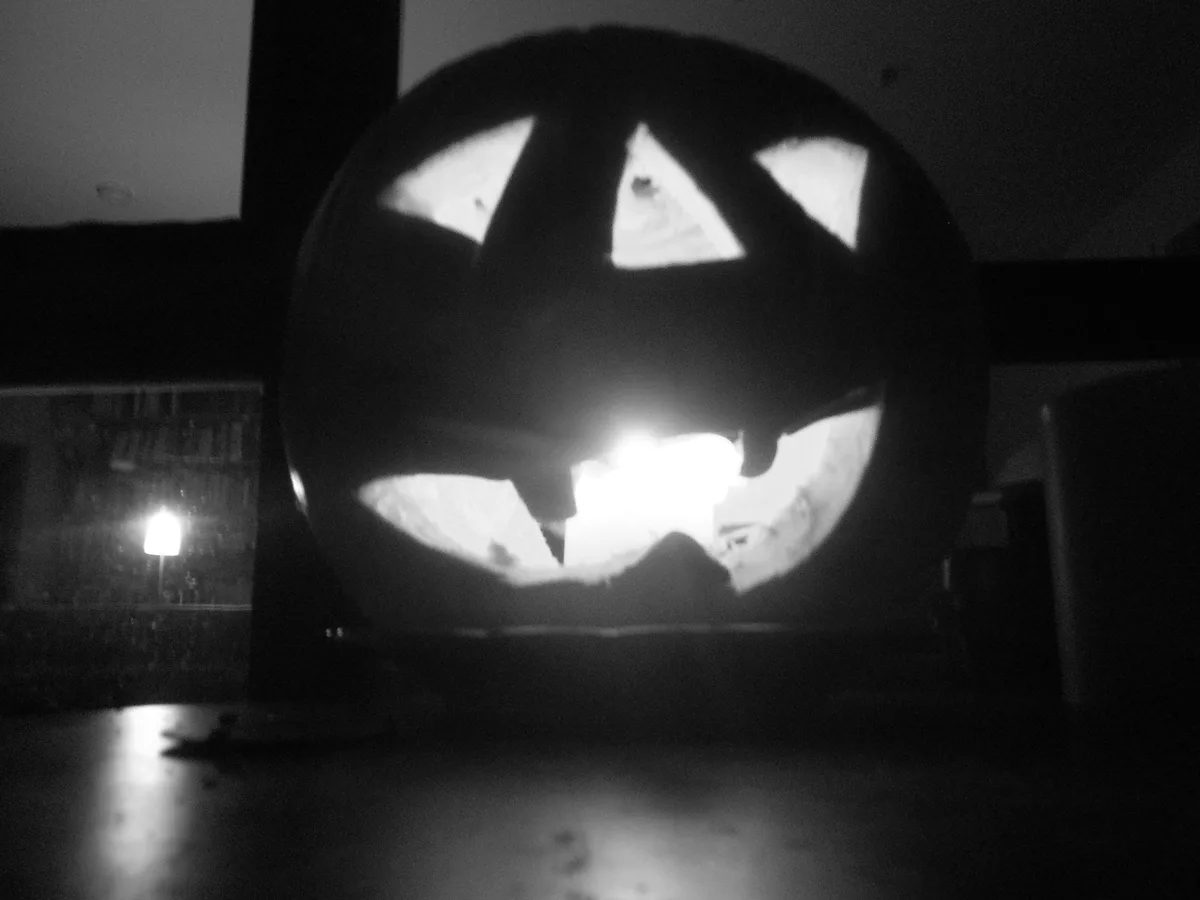 A black-and-white photo shows a carved pumpkin with triangular eyes and a wide, jagged mouth, illuminated from within. The…