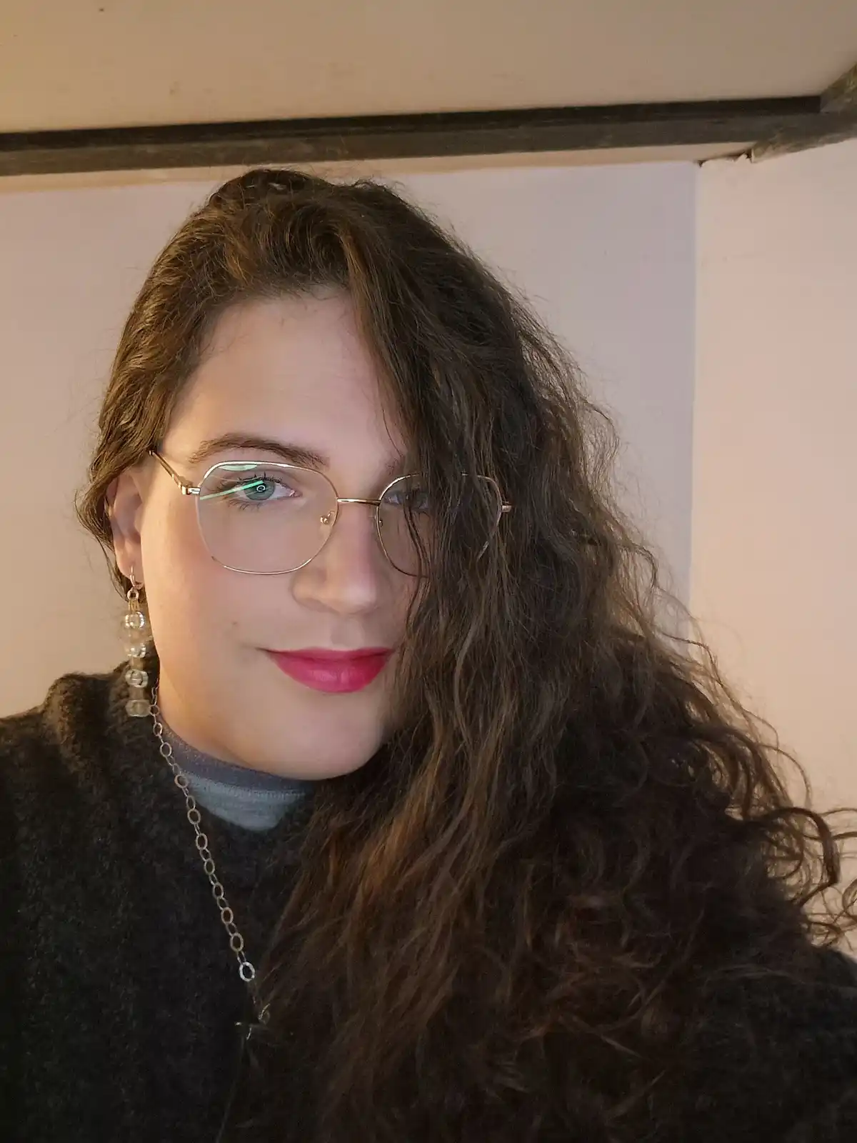 A person with long, wavy brown hair, wearing gold-rimmed glasses, a dark sweater over a grey turtleneck, and dangling…