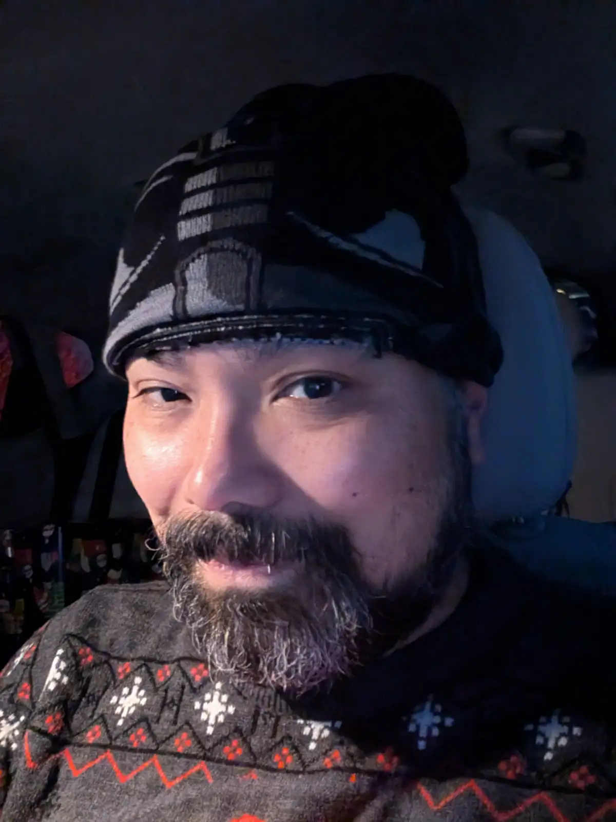 A man with a beard and a patterned beanie sits in a car, wearing a dark sweater with red and white geometric patterns. The…