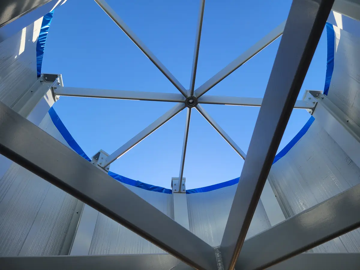 A low-angle photo looking up through the metal frame of a structure, possibly a dome or tower, at a clear blue sky. The…