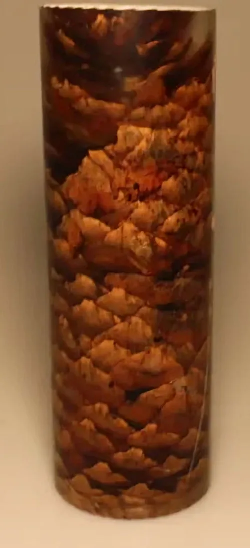 A cylindrical candle with a textured surface mimicking the layered, scaly pattern of a pinecone, featuring earthy brown and…