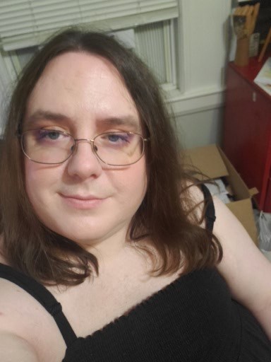 A person with glasses and dark hair looks at the camera with a tired expression, wearing a black top. Their eyes show…