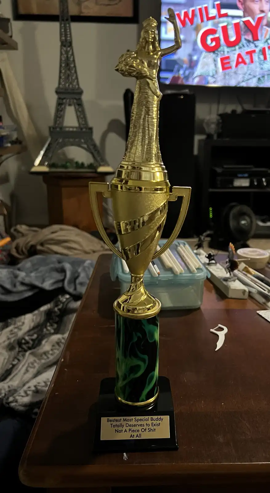 A golden trophy with a green marbled base sits on a wooden table, its plaque reading 'Bestest Most Special Buddy Totally…