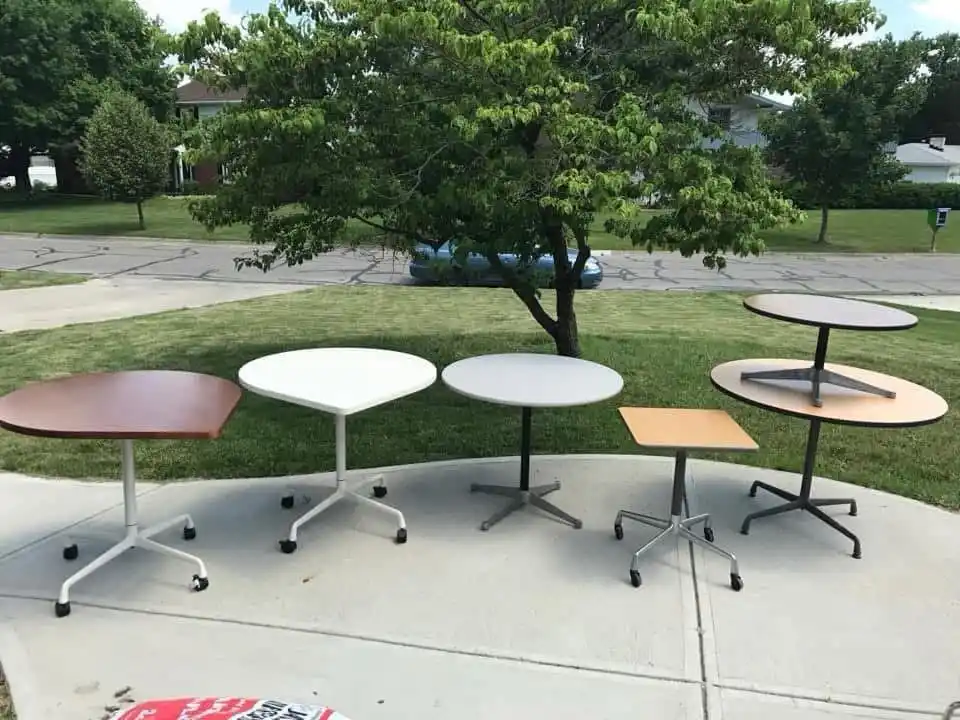Five small, wheeled office tables are arranged on a concrete sidewalk in a suburban yard, with a tree and street visible…
