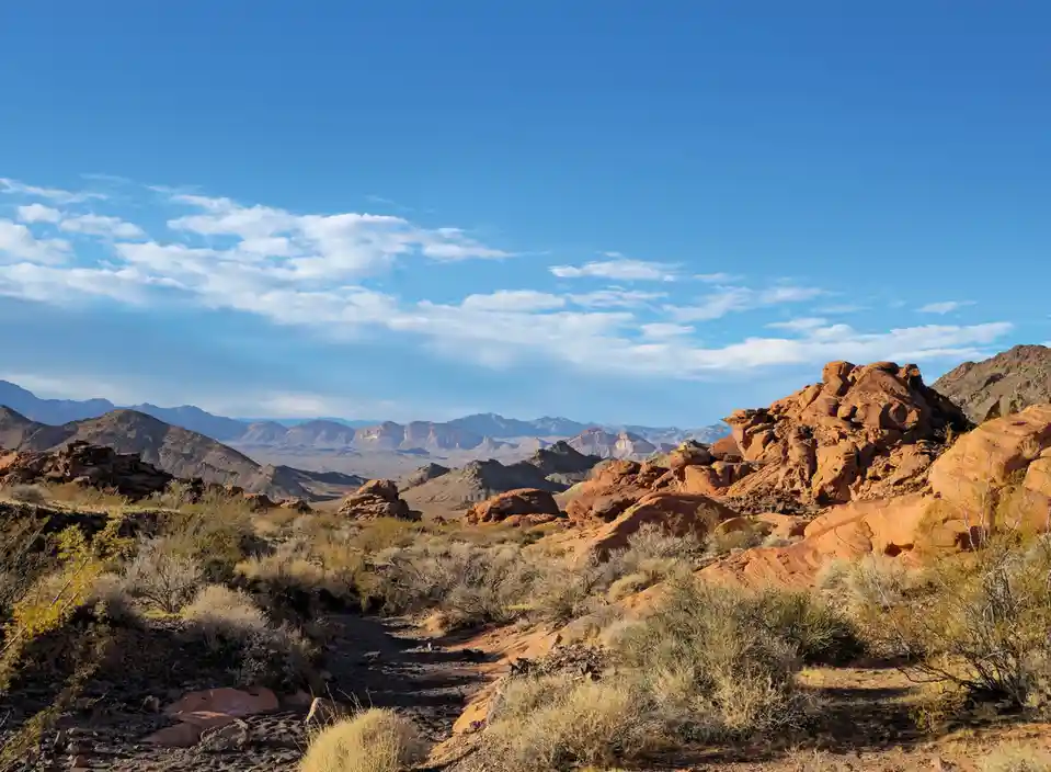 A scenic desert landscape with red rock formations, sparse shrubs, and distant mountains under a blue sky with scattered…