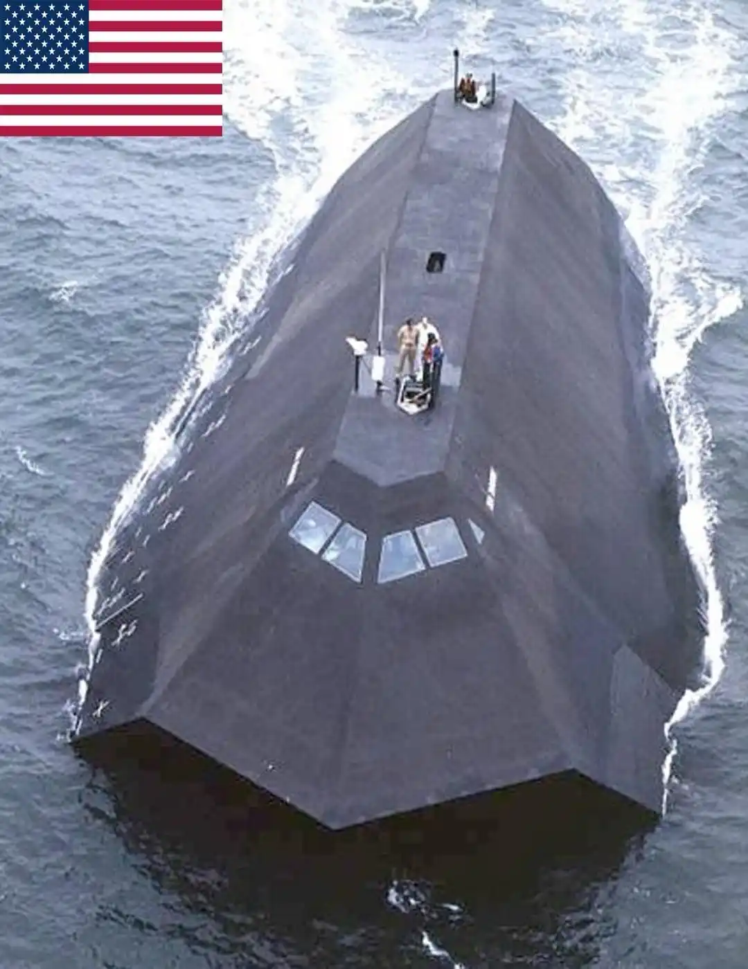 An aerial view of a dark gray, angular, stealth-like naval vessel cutting through choppy blue water, with several crew…