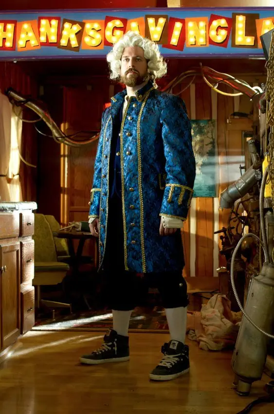 A man with a white wig and a blue ornate coat stands in a cluttered room with mismatched furniture and exposed pipes…