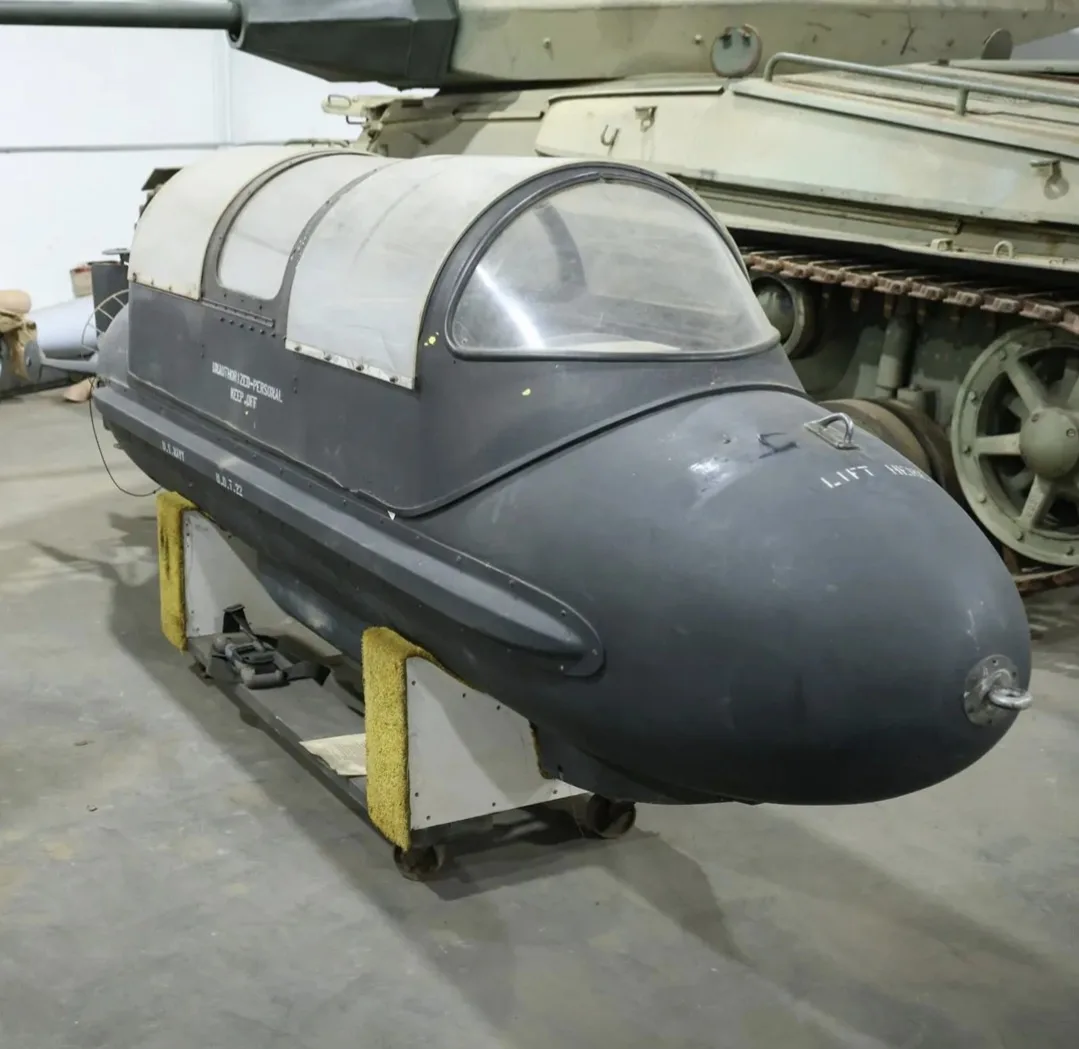 A black, egg-shaped, cockpit-equipped military device sits on a wheeled cart with yellow foam padding, displayed in a museum…
