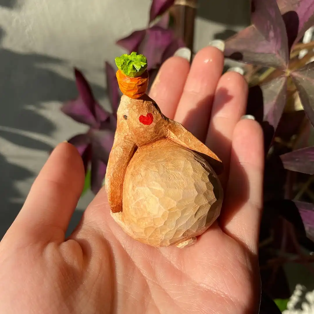 A hand holds a small, carved wooden rabbit figurine with a red heart painted on its cheek and a tiny carrot balanced on its…
