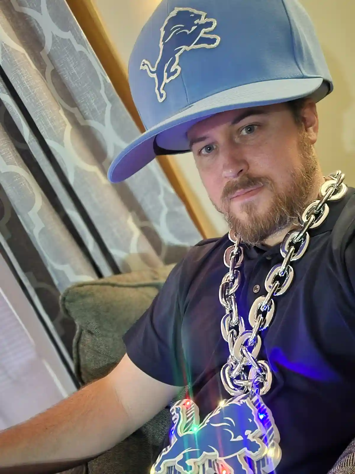 A man with a beard wears a blue Detroit Lions baseball cap and a large silver chain necklace with a glowing, multi-colored…