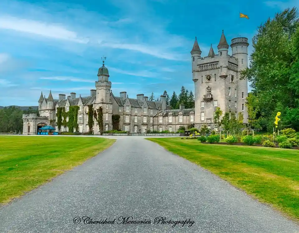 A grand, stone castle with turrets and ivy-covered walls stands at the end of a gravel driveway under a blue sky. The…