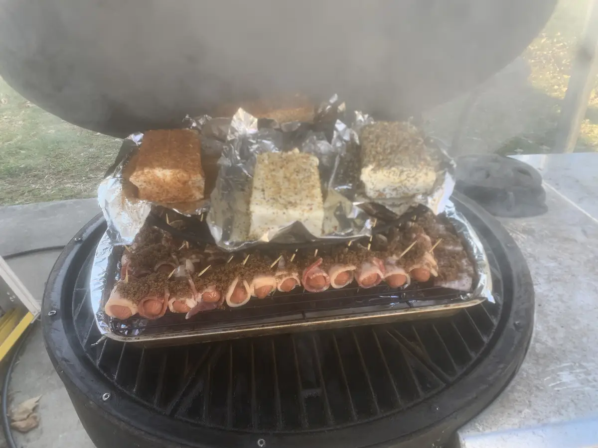 A black charcoal grill with its lid open, emitting smoke, contains two levels of food on foil-lined trays. The bottom tray…