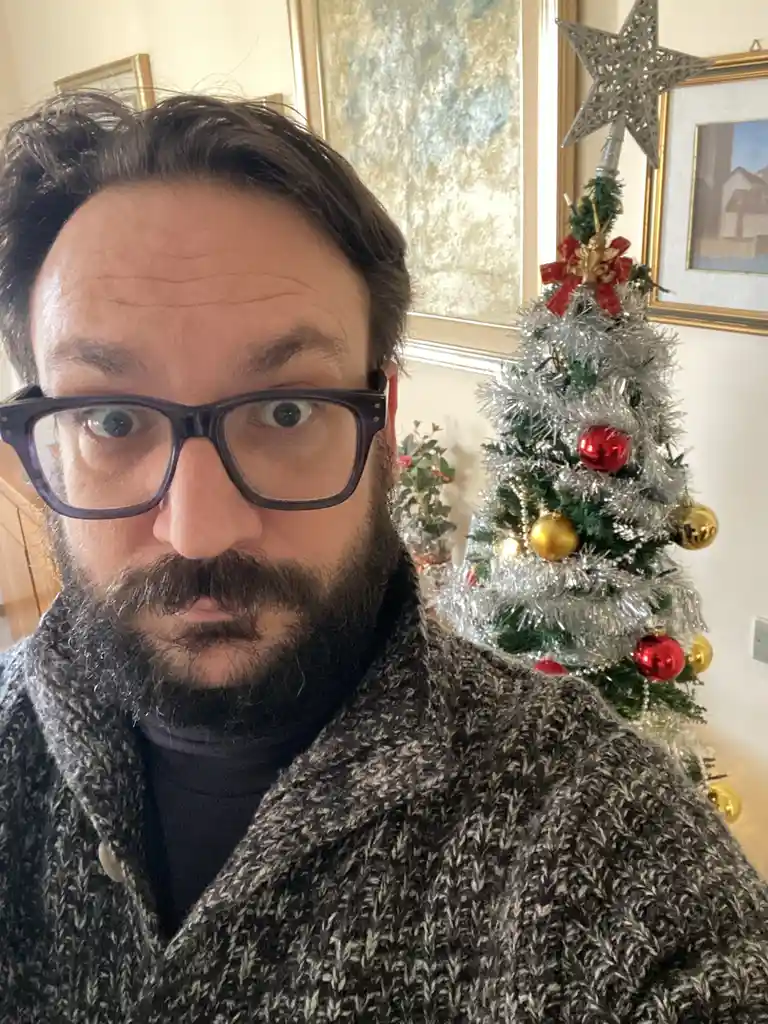A man with glasses and a beard stares directly at the camera with a wide-eyed, slightly horrified expression. Behind him, a…