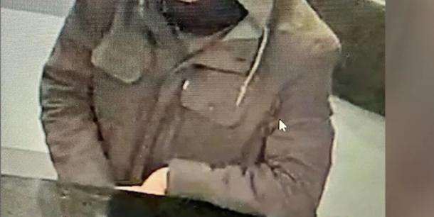 A low-resolution, grainy image shows a person wearing a brownish-gray jacket with a collar and pockets, standing indoors…