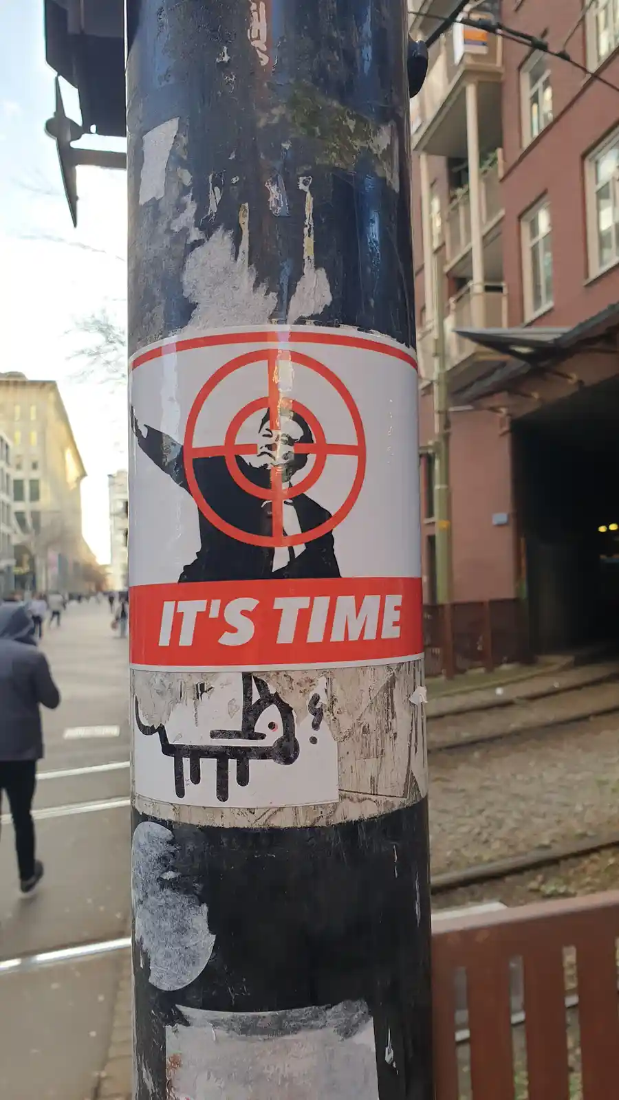 A street pole covered in peeling stickers features a prominent sticker showing a man in a suit with his arms raised…