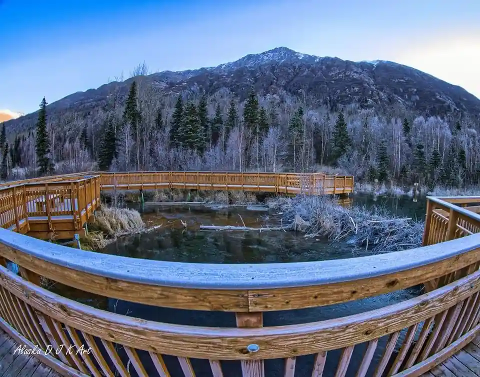 A scenic photograph shows a wooden boardwalk curving over a calm body of water, with a large, snow-dusted mountain rising…