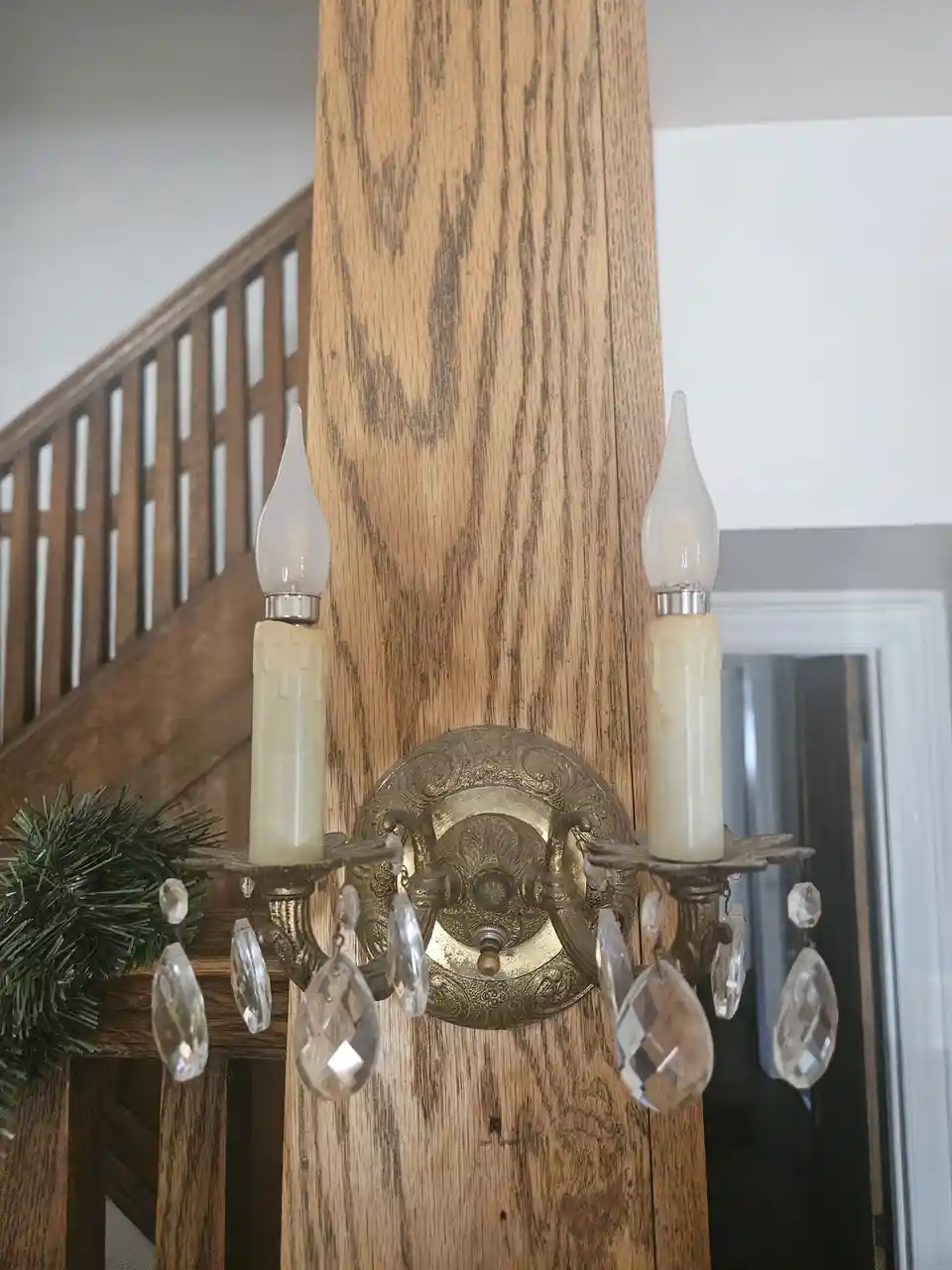 A vintage-style brass sconce with two candle-shaped LED bulbs and dangling crystal droplets is mounted on a thick wooden…