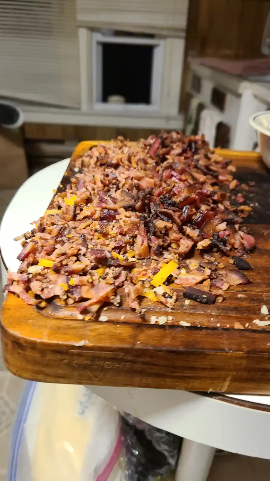 A wooden cutting board is covered in finely chopped, crispy bacon pieces mixed with small yellow and white fragments, likely…