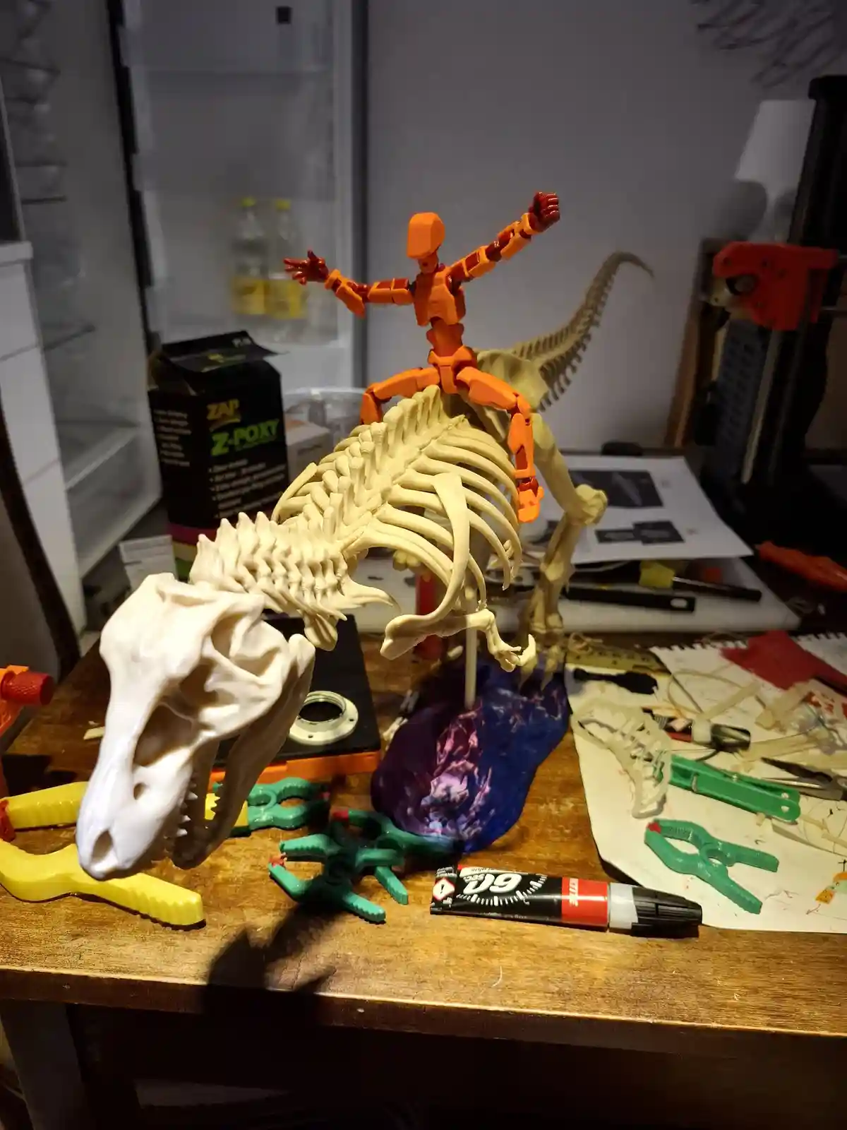 A 3D-printed dinosaur skeleton sits on a cluttered workbench surrounded by tools and glue, with an orange articulated robot…