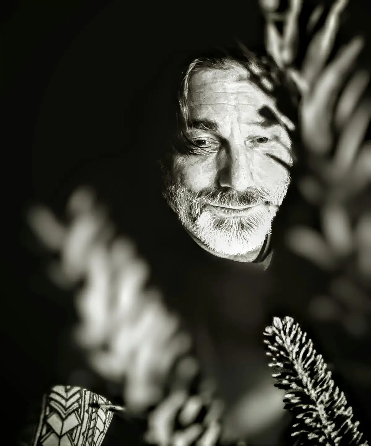 A black and white portrait of a smiling man with a beard, framed by out-of-focus foliage and branches. The text overlaid on…