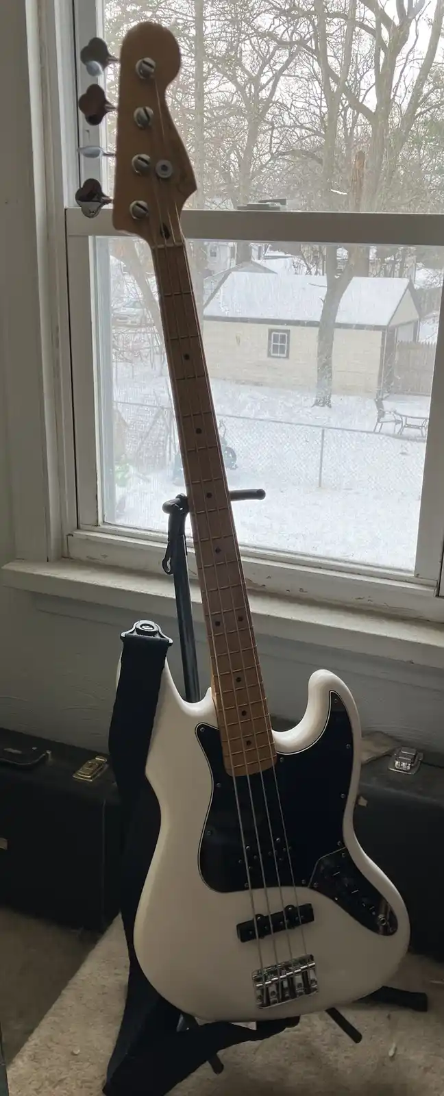 A white Fender-style bass guitar stands upright on a stand near a window, with a snowy outdoor scene visible through the…