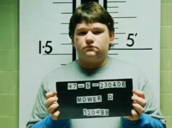 A young man with a serious expression stands in front of a height chart in a police station setting, holding a black mugshot…