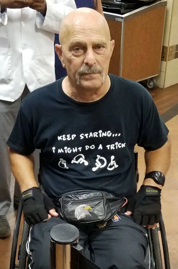A bald man with a mustache sits in a wheelchair wearing a black t-shirt with white text that reads 'KEEP STARING... I MIGHT…