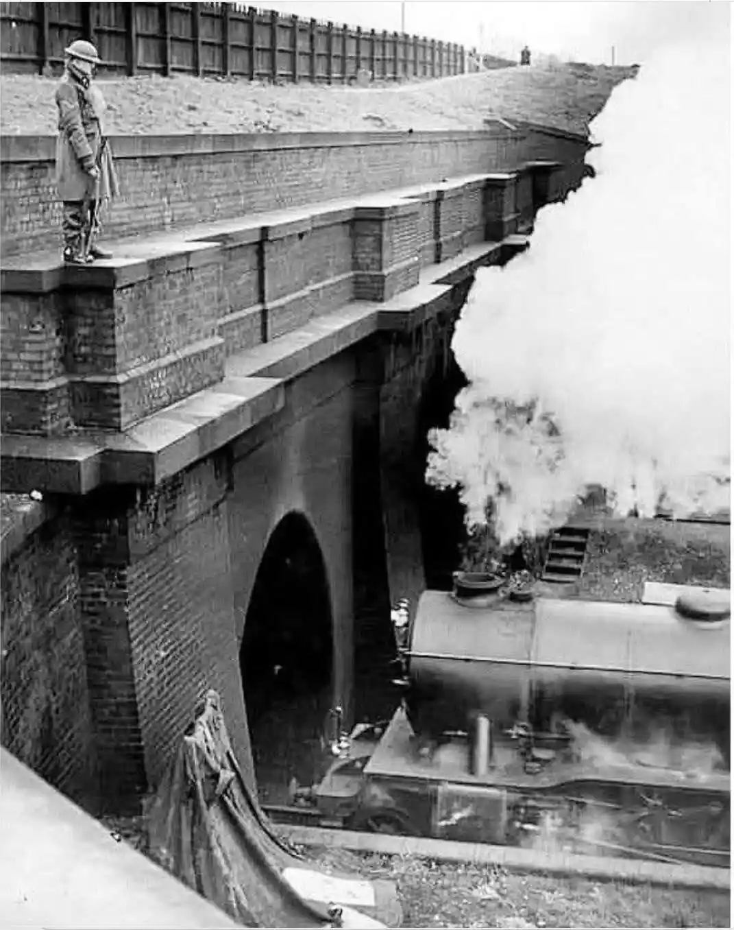 A black-and-white historical photo shows a soldier in uniform standing on a brick railway bridge, looking down at a steam…