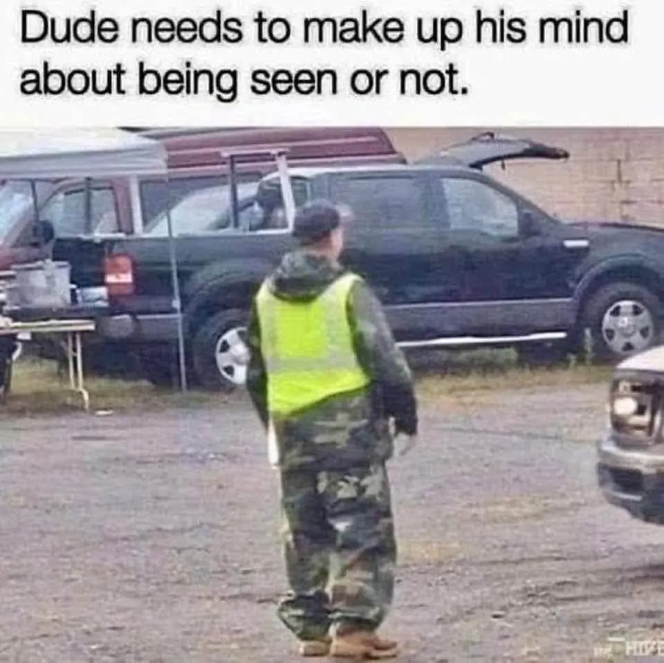 A person wearing a bright yellow safety vest over camouflage clothing stands with their back to the camera in a gravel lot…
