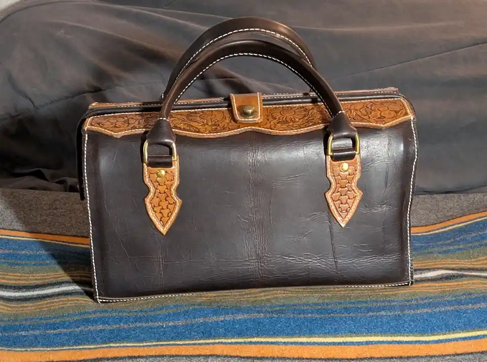 A dark brown leather handbag with tan tooled leather accents and white stitching sits on a striped blanket. The bag has a…