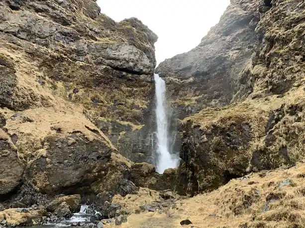 A scenic photo of a tall, narrow waterfall cascading down between two steep, moss-covered rock cliffs under an overcast sky…
