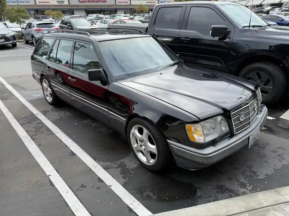 A black Mercedes-Benz station wagon with a red stripe parked in a wet parking lot, featuring a British flag emblem on its…