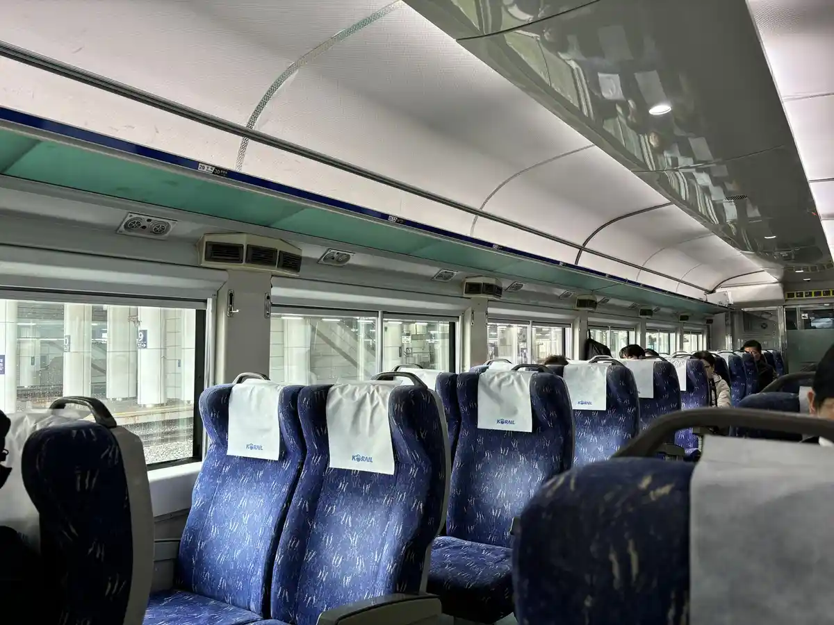 the image shows the interior of a korean train with blue patterned seats, white headrest covers with 'korail' branding, and…