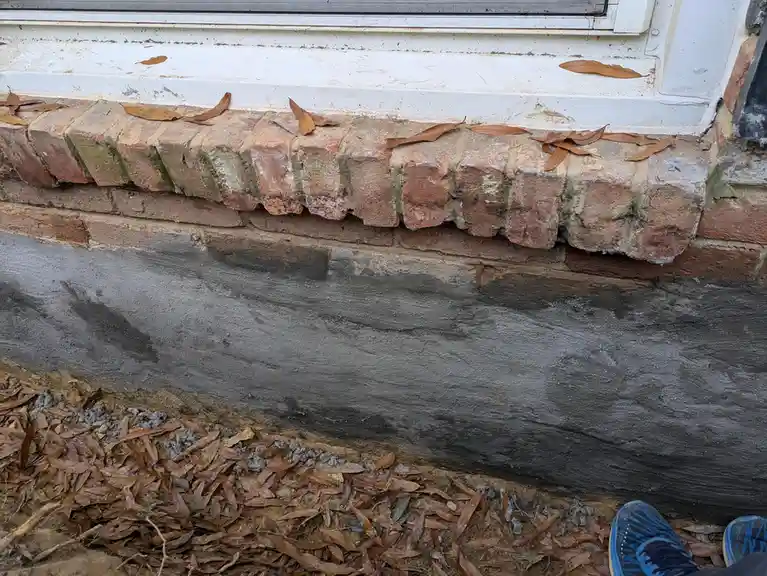 A close-up photo shows freshly poured wet concrete forming a new foundation or base along the bottom of a brick wall, with a…