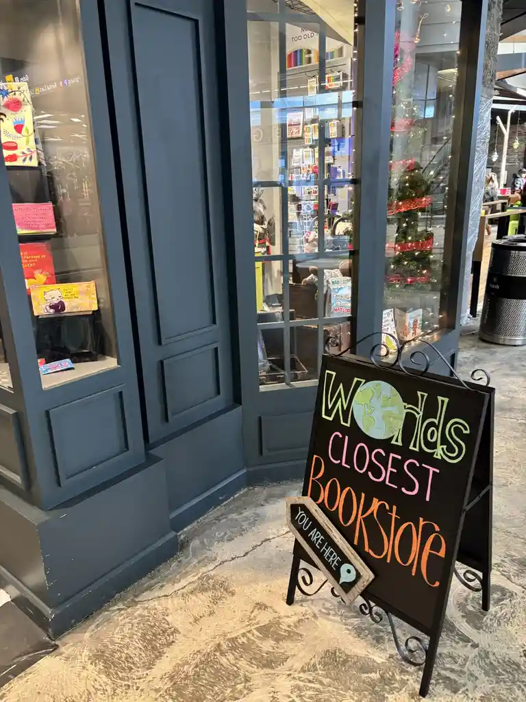 A black A-frame chalkboard sign outside a bookstore reads 'Worlds CLOSEST Bookstore' with 'Worlds' featuring a globe…
