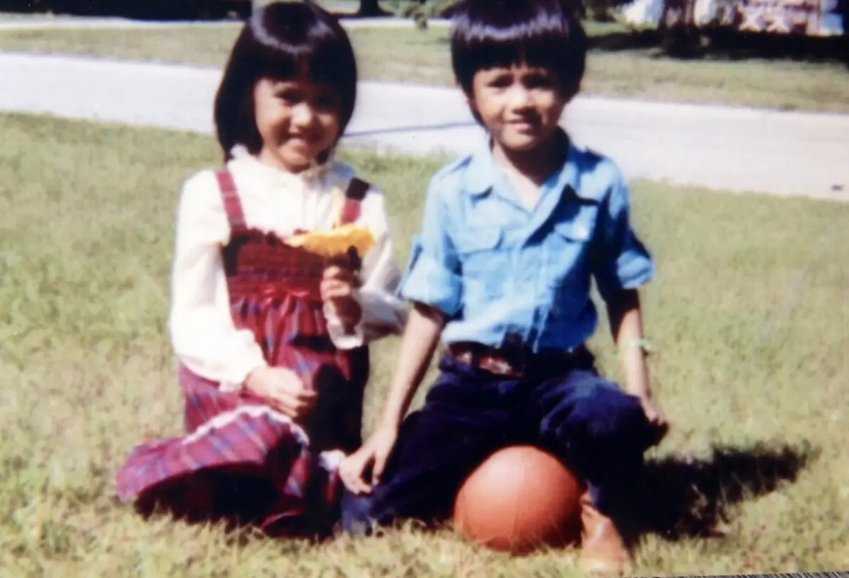 Two young children, a girl in a red plaid dress and a boy in a blue shirt, sit on grass next to a basketball. The girl holds…