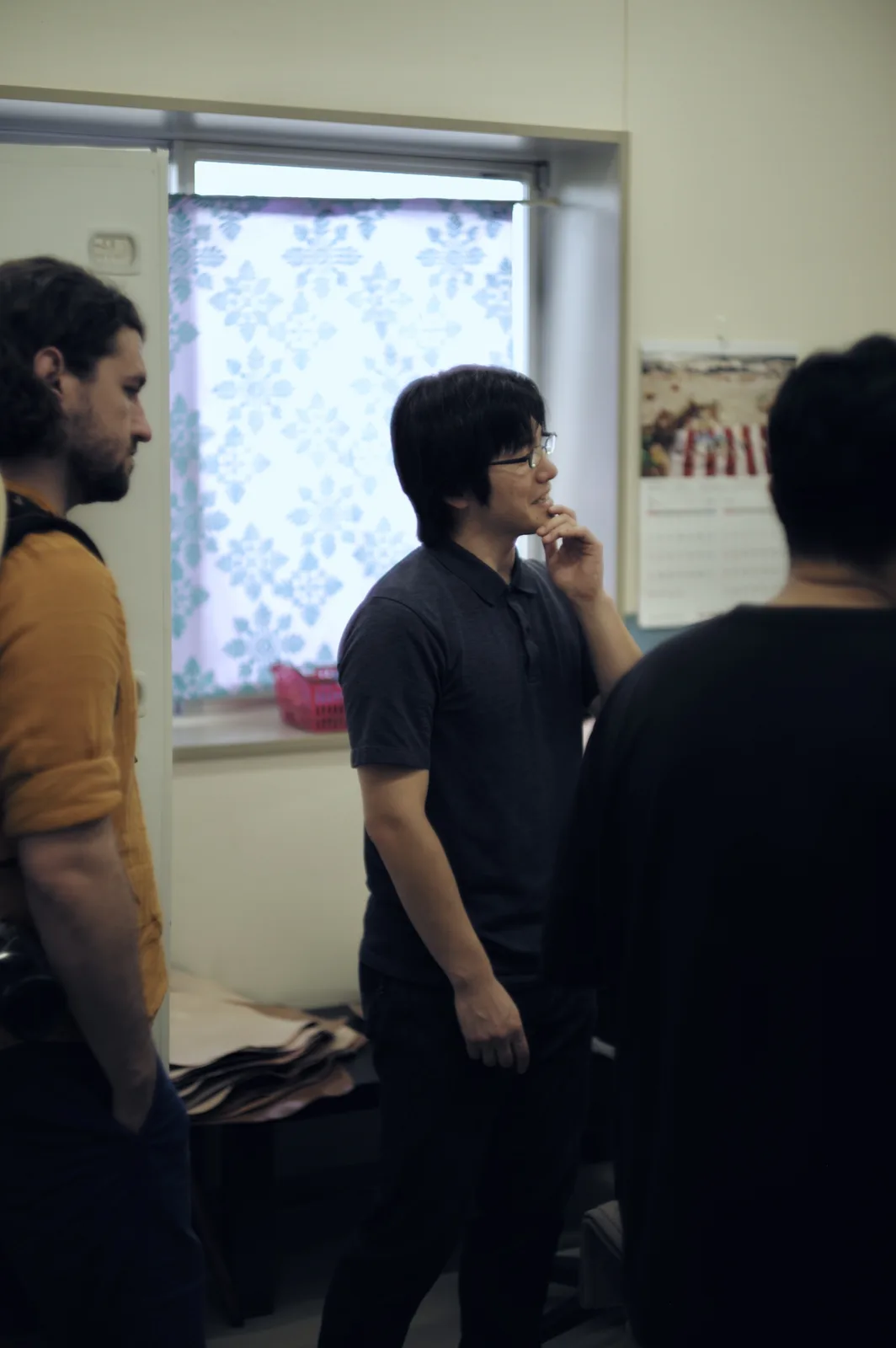 A man with glasses and dark hair, wearing a dark polo shirt, stands in a room looking thoughtful with his hand near his…