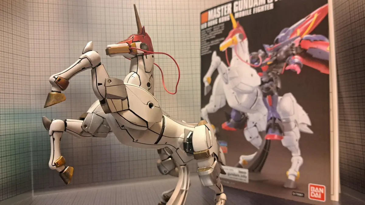 A white and gold mechanical unicorn model, resembling a Gundam, is posed rearing up in front of its box, which shows the…