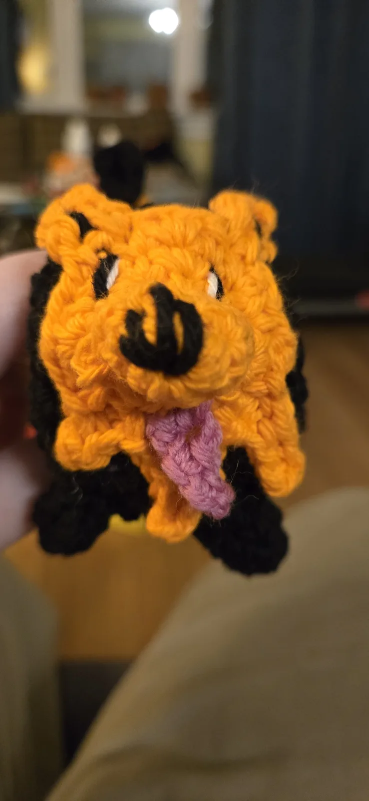 A close-up photo shows a handmade crocheted pig toy, primarily orange with black limbs and ears, holding a purple crocheted…