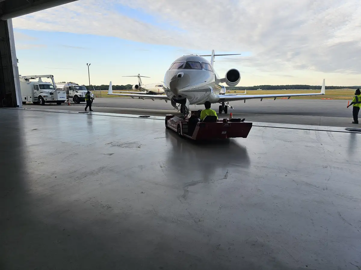 A large private jet is being pushed out of an airport hangar by a red tow tractor, with ground crew in yellow vests nearby…