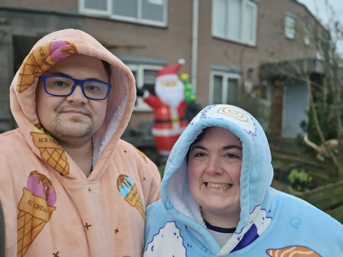 Two people stand outside in front of a house with a Santa inflatable in the background. The person on the left wears a…