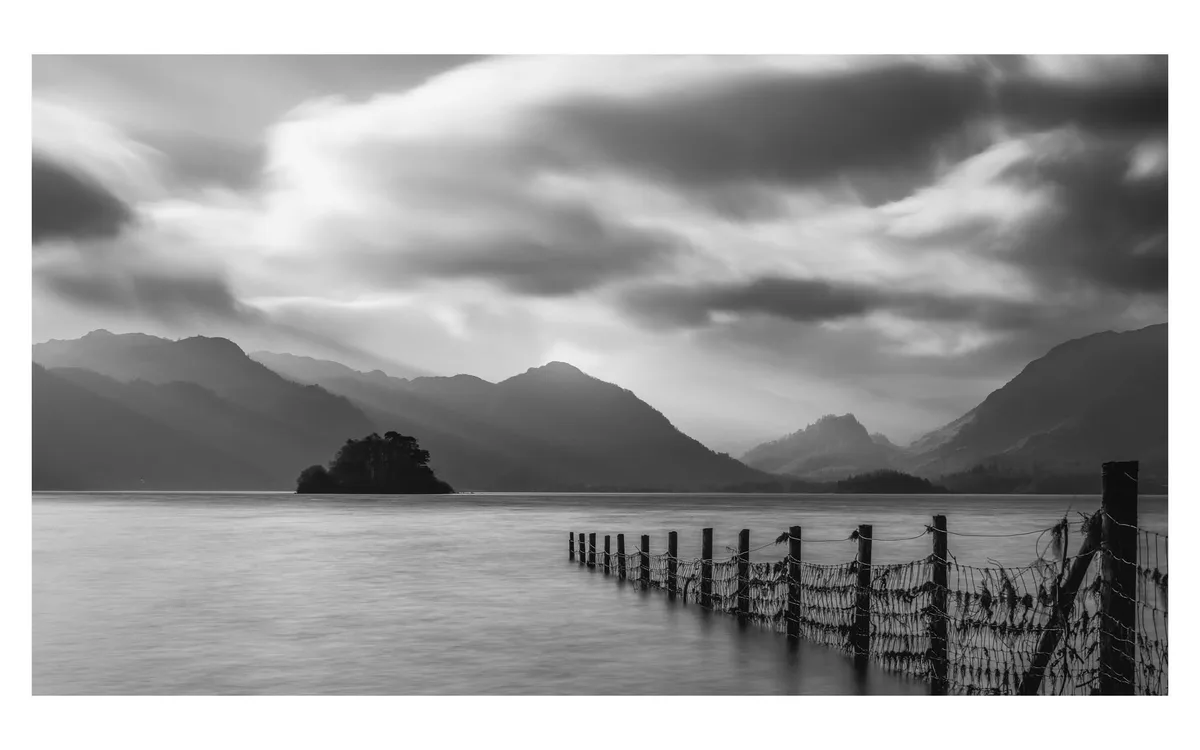 A black-and-white long-exposure photograph of a serene lake with misty mountains in the background and a wooden fence…