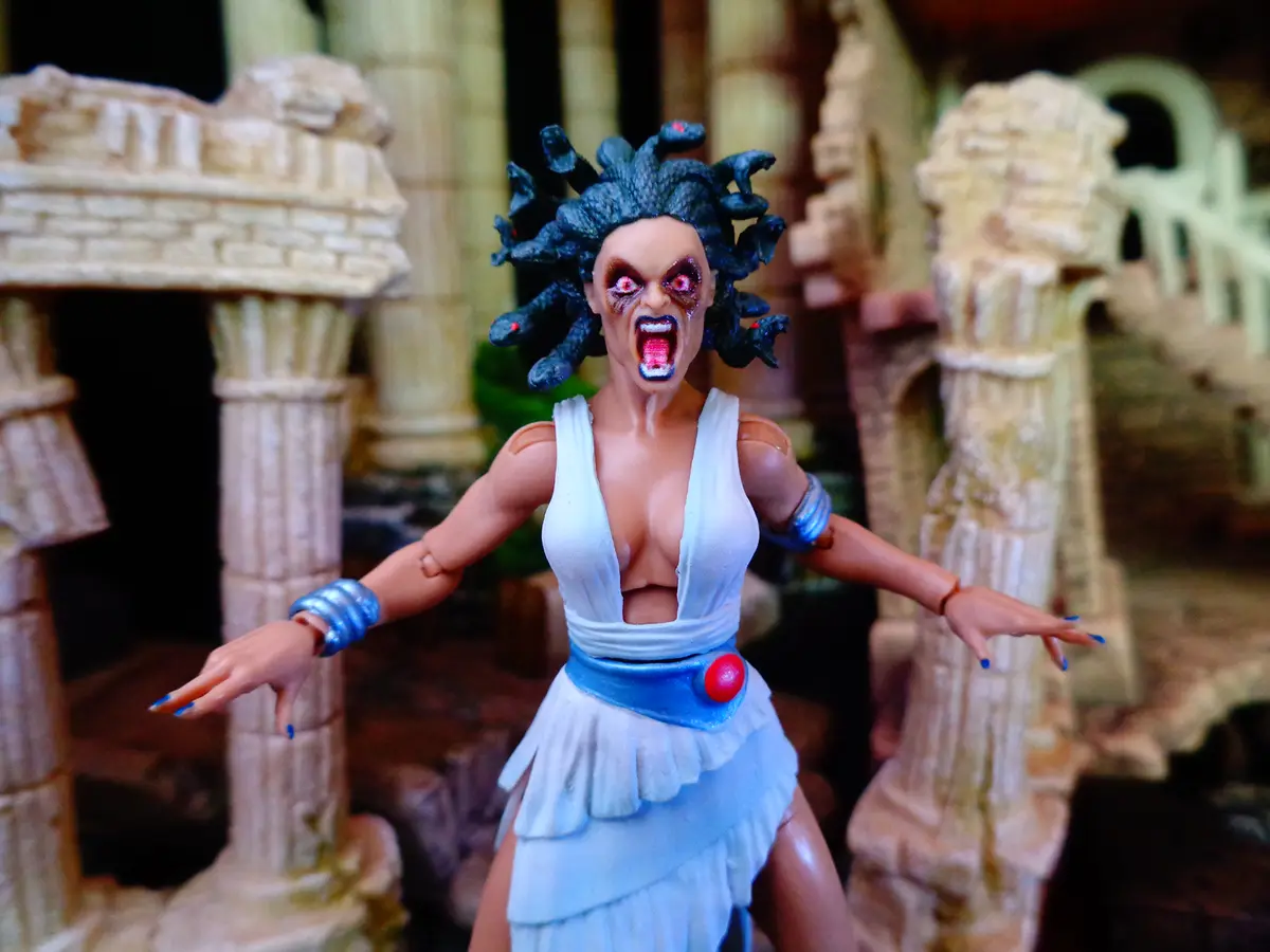 A Medusa action figure with a screaming, angry expression, wearing a white dress and blue accessories, stands with arms…