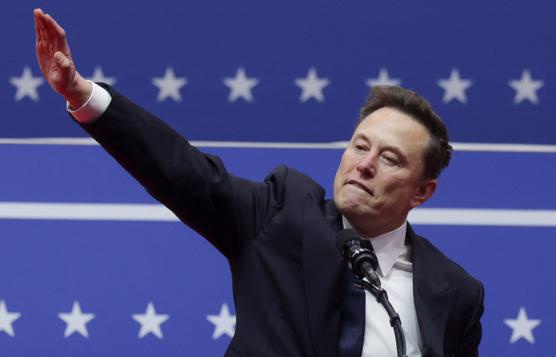 elon musk stands at a podium in front of a blue backdrop with white stars and stripes, raising his right hand in a waving…
