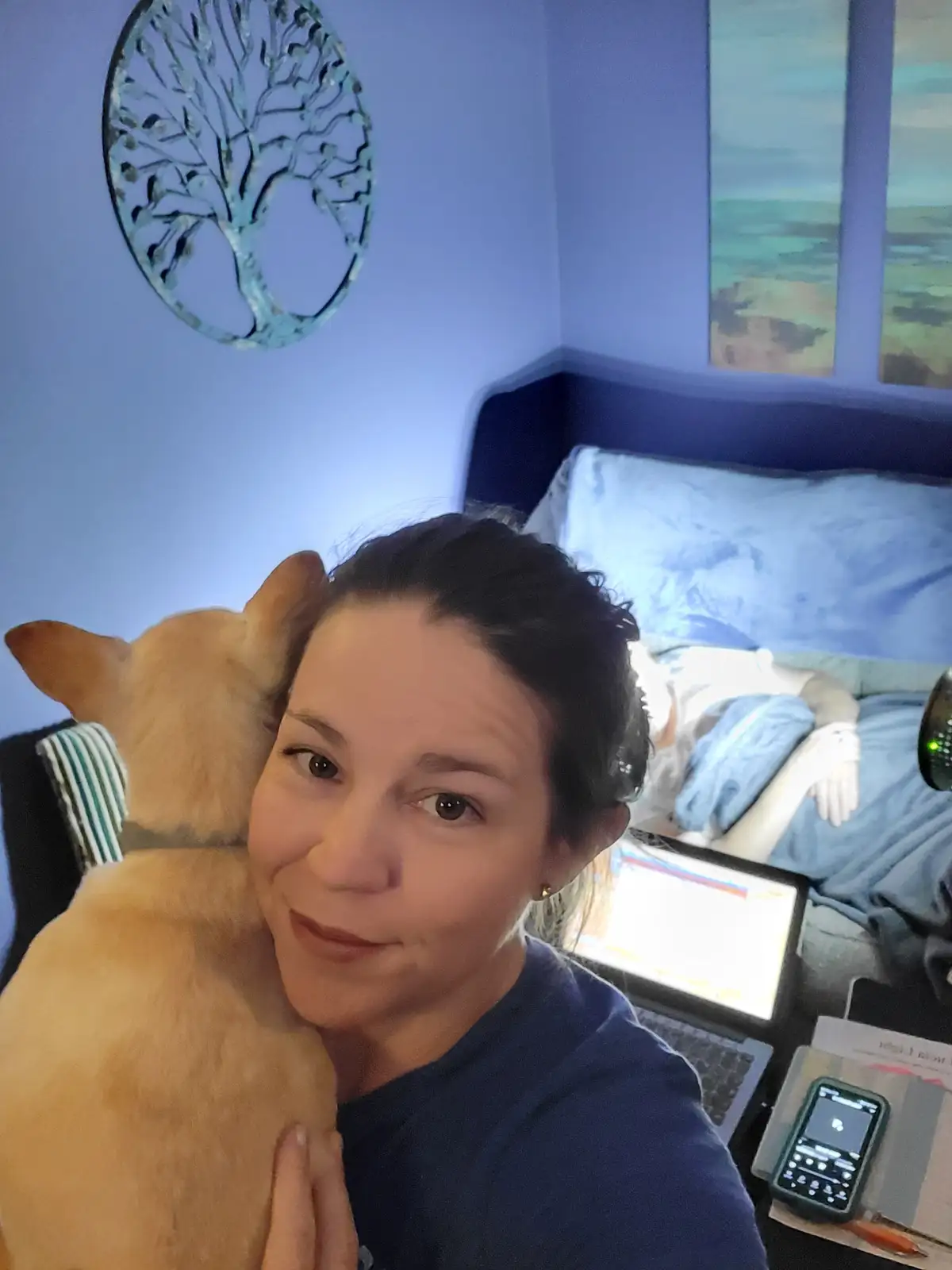 A woman takes a selfie in her bedroom, smiling while a light brown dog rests its head on her shoulder. In the background…