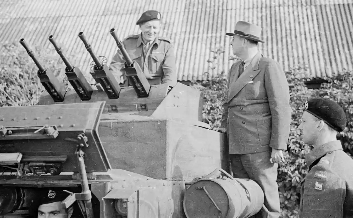 A black-and-white historical photo shows a man in a suit standing beside a military tank with multiple machine guns, smiling…