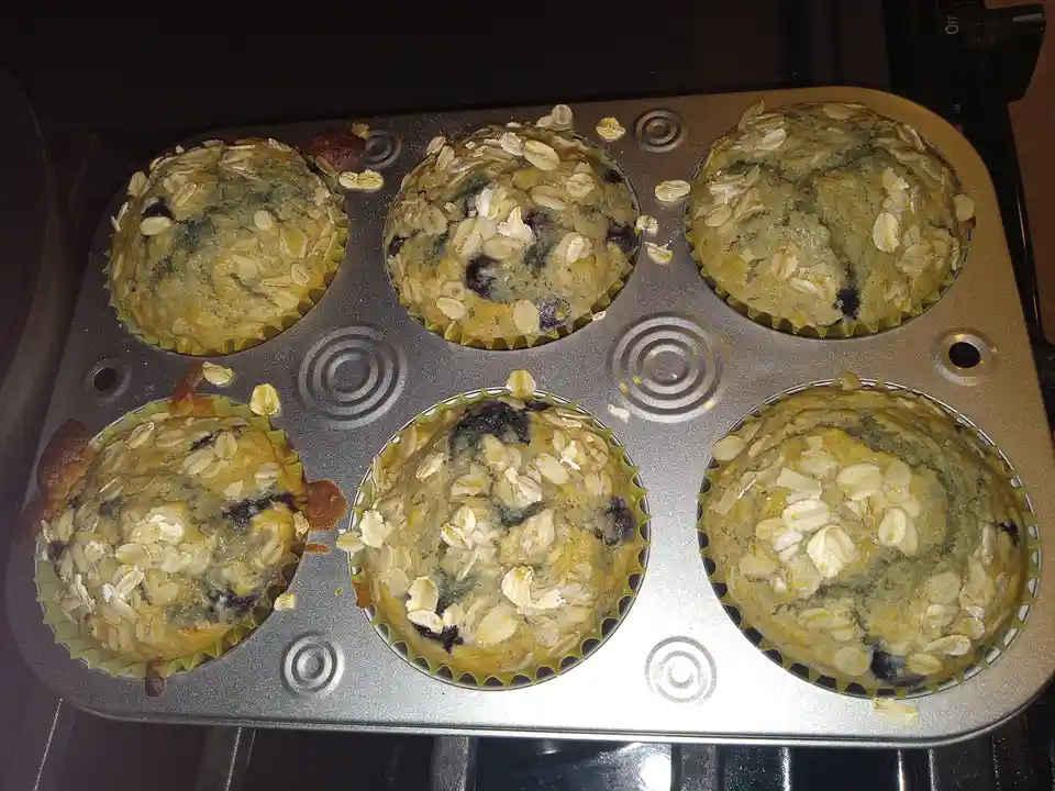 Six freshly baked blueberry oat muffins in a metal muffin tin, some slightly overflowing and spilling oats. The text overlay…