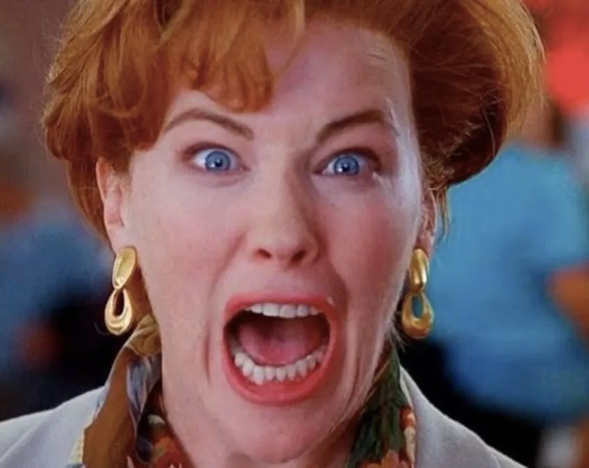 A close-up of a woman with red hair and blue eyes, wearing large gold hoop earrings and a patterned scarf, screaming with…