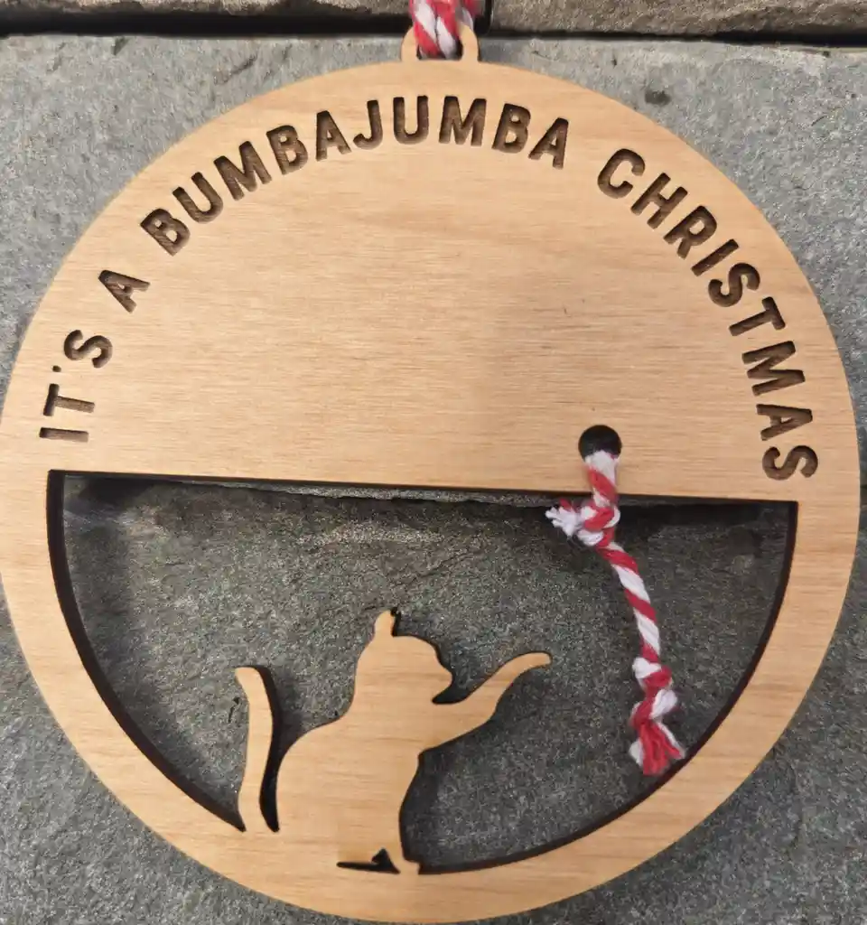 A circular wooden ornament with engraved text reading 'IT'S A BUMBAJUMBA CHRISTMAS' around the top half, and a cutout…